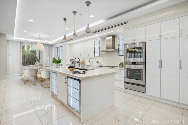 kitchen featuring wall chimney exhaust hood, a tray ceiling, stainless steel double oven, white cabinetry, and a warming drawer