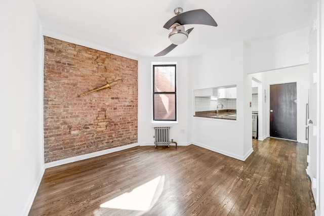 unfurnished living room with radiator, brick wall, baseboards, dark wood finished floors, and a ceiling fan