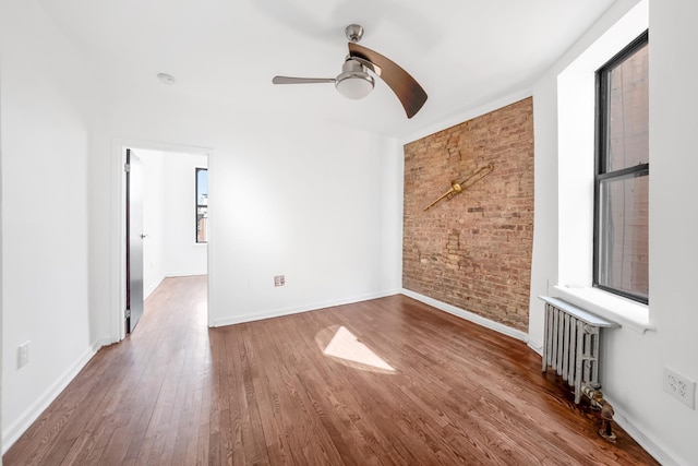 unfurnished room featuring radiator, wood finished floors, baseboards, brick wall, and ceiling fan