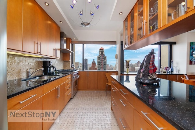 kitchen with glass insert cabinets, high end range, dark stone countertops, wall chimney range hood, and a sink