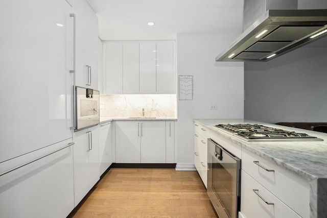 kitchen with wall chimney range hood, stainless steel gas cooktop, light wood-style flooring, white cabinets, and modern cabinets