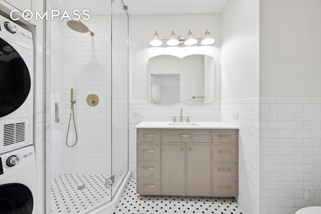 bathroom featuring wainscoting, a tile shower, vanity, stacked washing maching and dryer, and tile walls