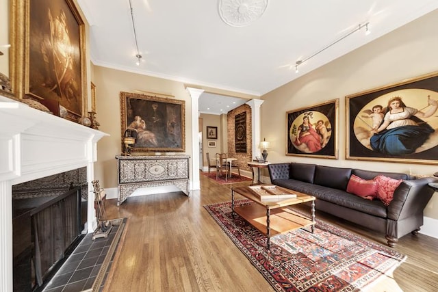 living area with wood finished floors, rail lighting, a fireplace, crown molding, and decorative columns