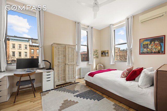 bedroom with radiator, baseboards, an AC wall unit, light wood-style floors, and a ceiling fan