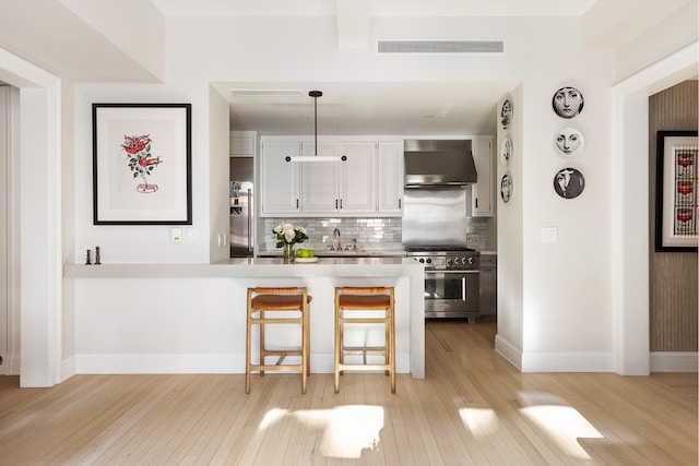 kitchen featuring wall chimney range hood, decorative backsplash, high end stainless steel range, a peninsula, and light wood-style flooring