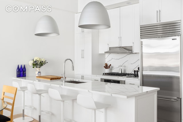 kitchen featuring light stone counters, a sink, under cabinet range hood, appliances with stainless steel finishes, and tasteful backsplash