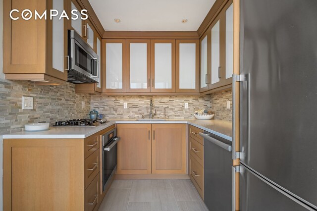 kitchen featuring stainless steel appliances, a sink, light countertops, tasteful backsplash, and glass insert cabinets