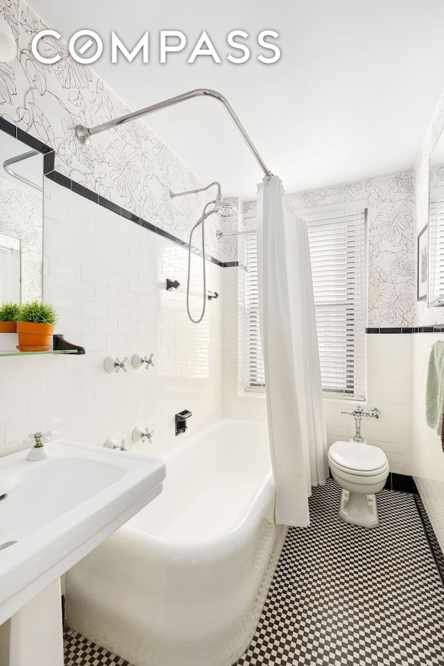 bathroom with shower / bath combo with shower curtain, toilet, tile walls, wainscoting, and wallpapered walls