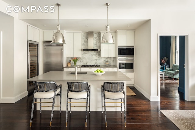 kitchen featuring built in appliances, a sink, white cabinetry, decorative backsplash, and wall chimney exhaust hood