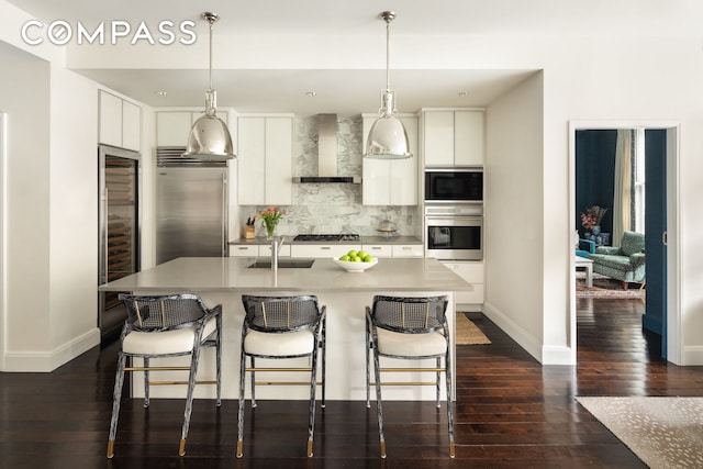 kitchen featuring built in appliances, wall chimney exhaust hood, a sink, tasteful backsplash, and white cabinets