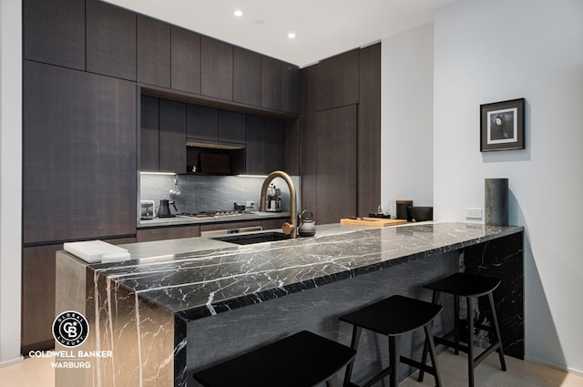 kitchen featuring tasteful backsplash, dark stone counters, a kitchen breakfast bar, dark brown cabinets, and a sink