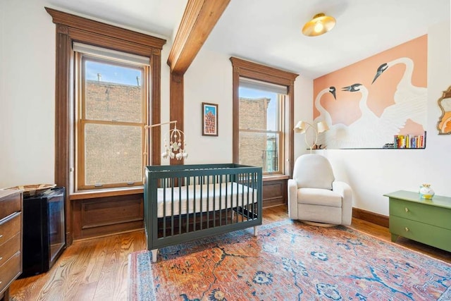 bedroom featuring a nursery area, beam ceiling, baseboards, and wood finished floors