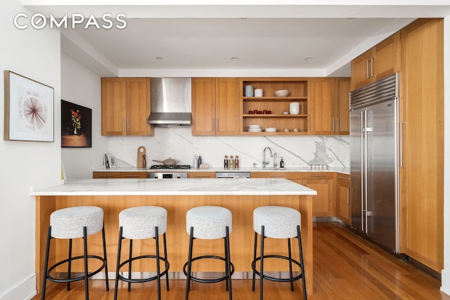 kitchen with open shelves, appliances with stainless steel finishes, brown cabinetry, a peninsula, and wall chimney exhaust hood