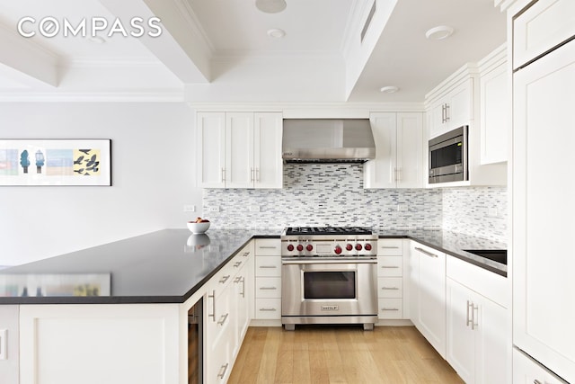 kitchen featuring a peninsula, appliances with stainless steel finishes, wall chimney exhaust hood, dark countertops, and a raised ceiling