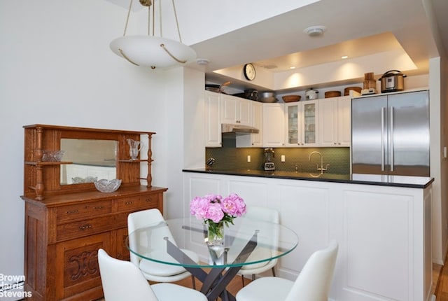 kitchen featuring built in fridge, dark countertops, decorative backsplash, glass insert cabinets, and under cabinet range hood