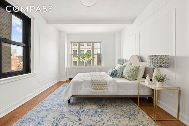 bedroom featuring beamed ceiling, a decorative wall, baseboards, and wood finished floors