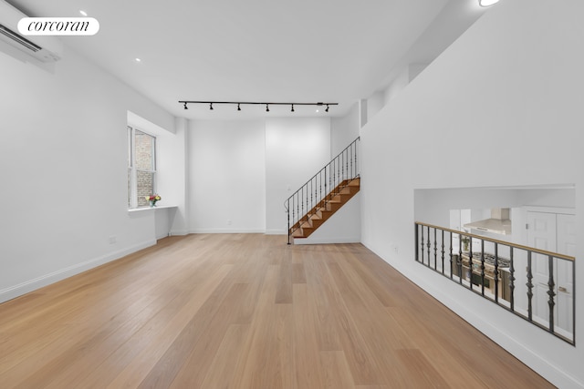 unfurnished living room with wood finished floors, a wall unit AC, rail lighting, baseboards, and stairs