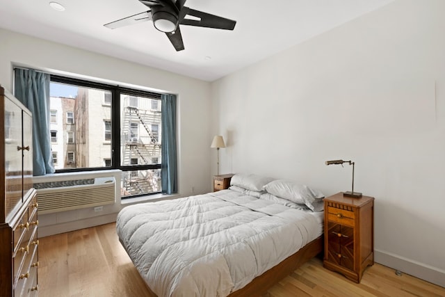 bedroom featuring a wall unit AC, baseboards, light wood finished floors, and ceiling fan