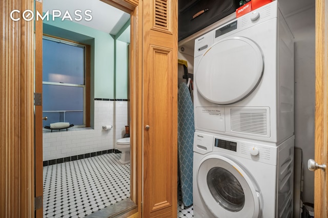 clothes washing area featuring tile patterned floors, tile walls, wainscoting, stacked washer / dryer, and laundry area