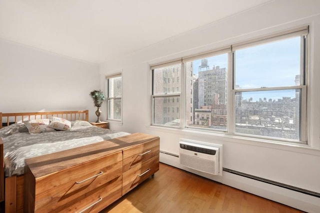 bedroom with a wall mounted air conditioner, light wood-type flooring, a city view, and a baseboard radiator