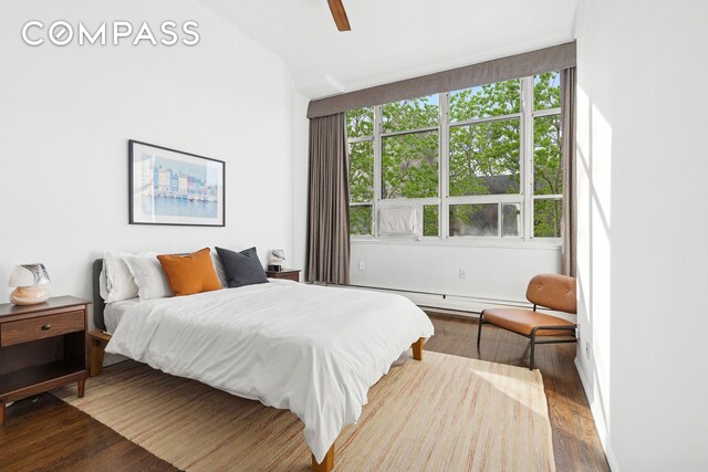 bedroom with wood finished floors, multiple windows, a baseboard heating unit, and ceiling fan