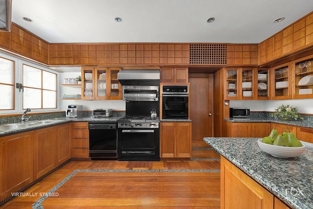 kitchen with black appliances, a sink, wall chimney exhaust hood, brown cabinetry, and glass insert cabinets