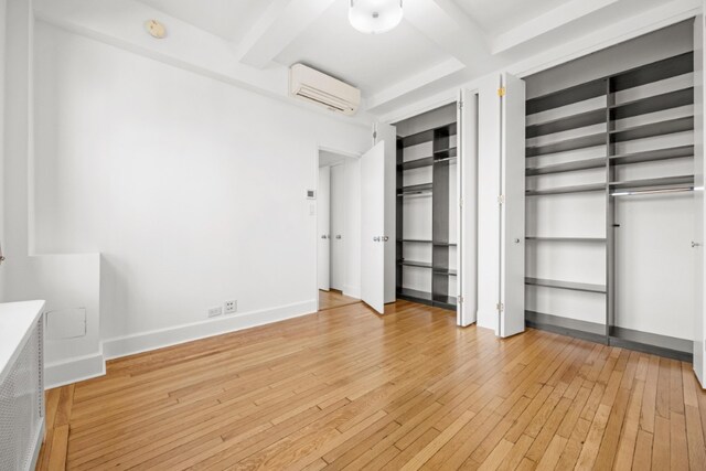 unfurnished bedroom featuring multiple closets, beam ceiling, baseboards, an AC wall unit, and light wood-style flooring