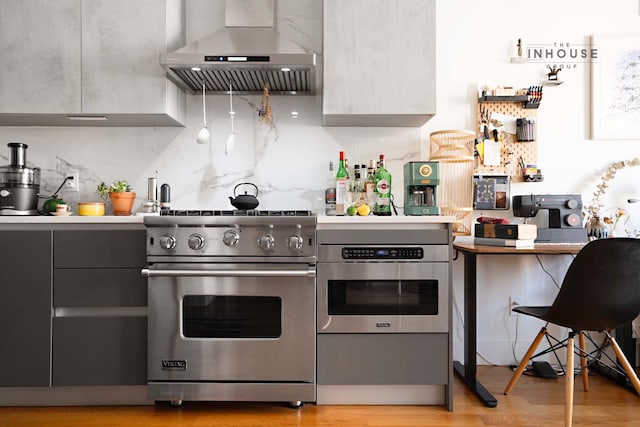 kitchen with light wood finished floors, decorative backsplash, gray cabinets, stainless steel appliances, and wall chimney exhaust hood