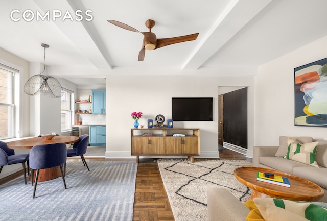 living room with baseboards, beam ceiling, beverage cooler, and ceiling fan with notable chandelier