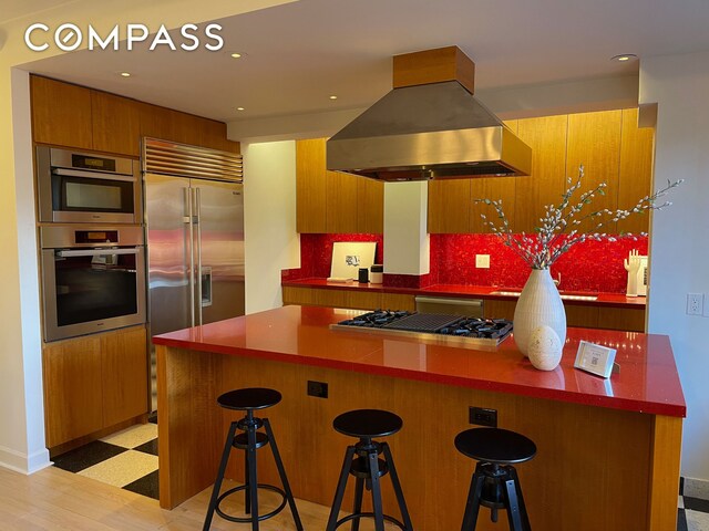 227 E 57th St Unit 8Efg, New York City, NY 10022 - kitchen with island range hood, appliances with stainless steel finishes, brown cabinets, light floors, and a breakfast bar