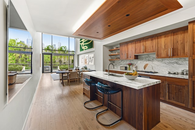 kitchen featuring open shelves, light wood-style flooring, a kitchen island with sink, a sink, and backsplash