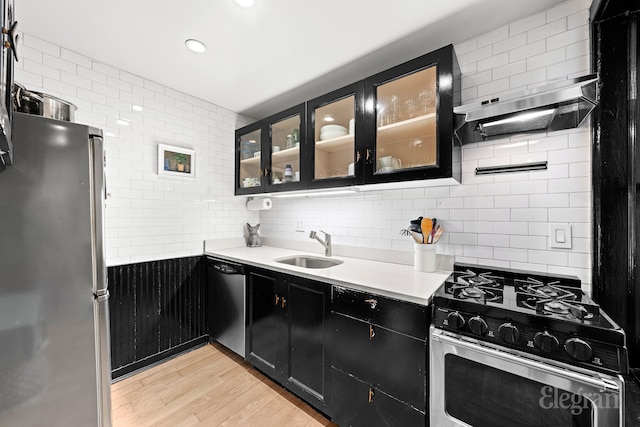 kitchen featuring light wood finished floors, a sink, stainless steel appliances, wall chimney exhaust hood, and dark cabinets