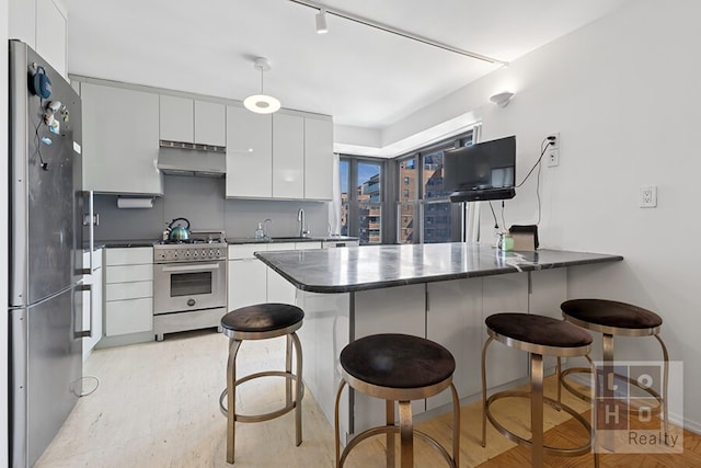 kitchen featuring a peninsula, stainless steel appliances, under cabinet range hood, dark countertops, and a kitchen breakfast bar