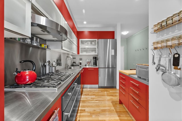 kitchen with a sink, stainless steel appliances, light wood-style floors, stainless steel counters, and wall chimney exhaust hood