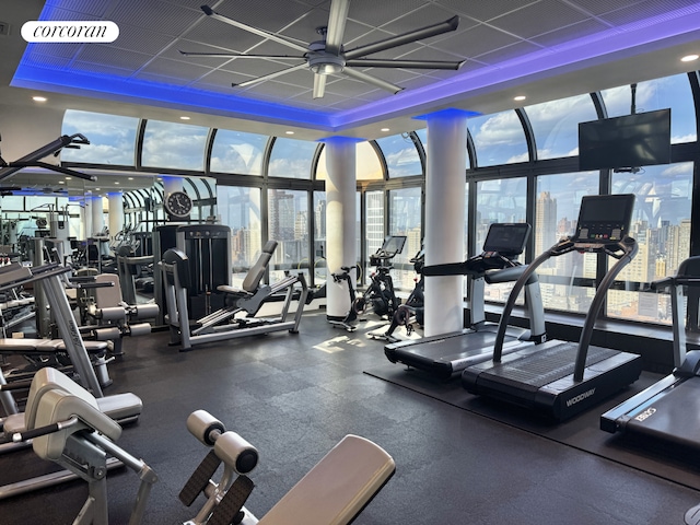 gym with plenty of natural light, a ceiling fan, a raised ceiling, and a wall of windows