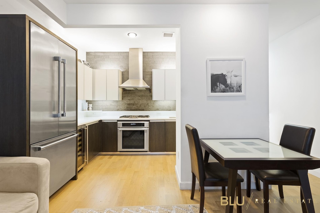 kitchen featuring light wood-type flooring, white cabinetry, appliances with stainless steel finishes, wall chimney exhaust hood, and light countertops