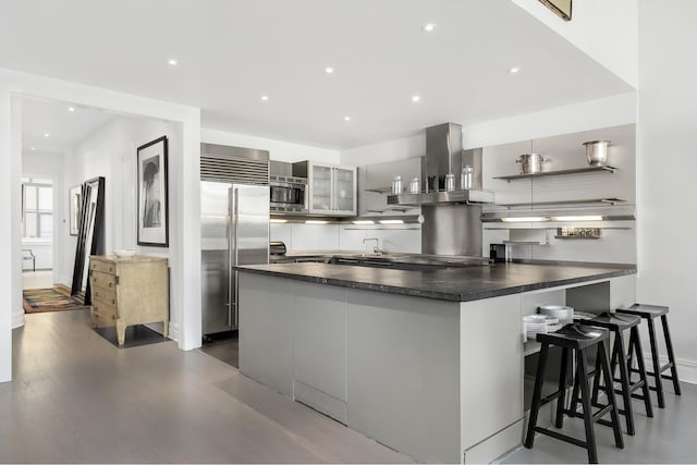 kitchen featuring dark countertops, built in appliances, dark wood finished floors, a peninsula, and island exhaust hood