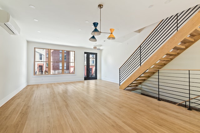 unfurnished living room featuring a notable chandelier, a wall mounted AC, wood finished floors, baseboards, and stairs