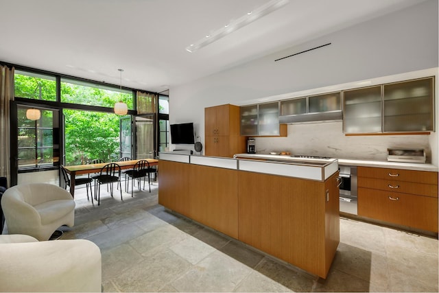 kitchen featuring stone tile floors, light countertops, glass insert cabinets, under cabinet range hood, and tasteful backsplash