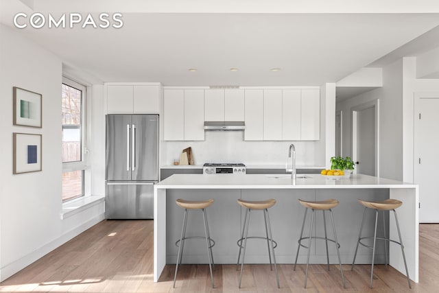 kitchen featuring a sink, light countertops, high end refrigerator, white cabinetry, and under cabinet range hood