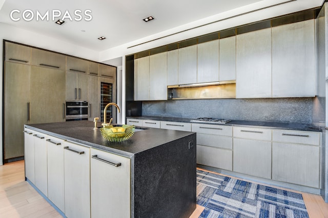 kitchen featuring dark countertops, tasteful backsplash, a center island with sink, light wood-style flooring, and appliances with stainless steel finishes