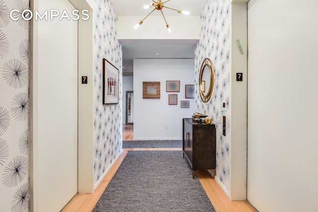 corridor with wood finished floors, elevator, wallpapered walls, baseboards, and a chandelier