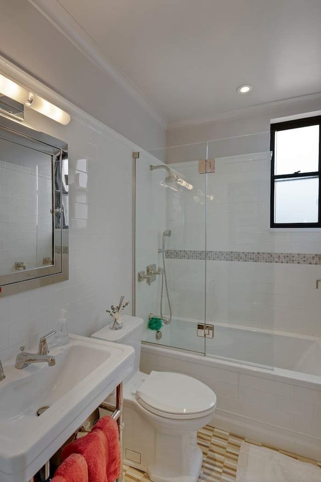 bathroom featuring toilet, ornamental molding, combined bath / shower with glass door, tile walls, and a sink