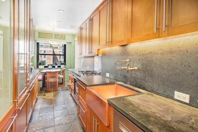 kitchen featuring dark stone countertops, brown cabinetry, stainless steel gas cooktop, stone tile flooring, and tasteful backsplash