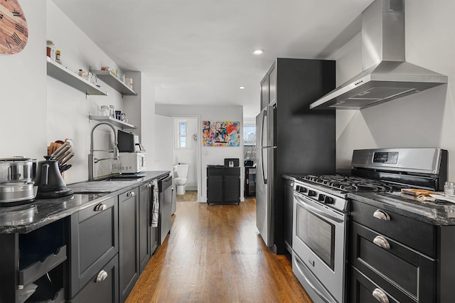 kitchen featuring open shelves, wood finished floors, stainless steel appliances, wall chimney exhaust hood, and dark cabinets