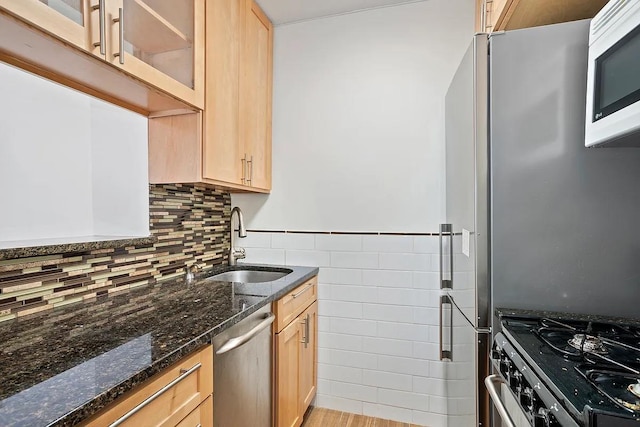 kitchen with stainless steel appliances, a sink, light brown cabinetry, glass insert cabinets, and dark stone countertops