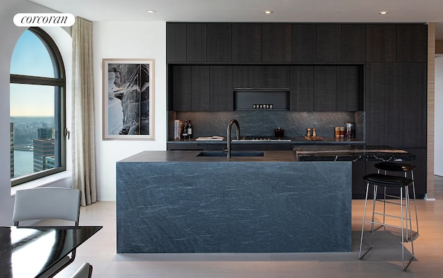 kitchen featuring dark cabinets, an island with sink, decorative backsplash, a sink, and modern cabinets