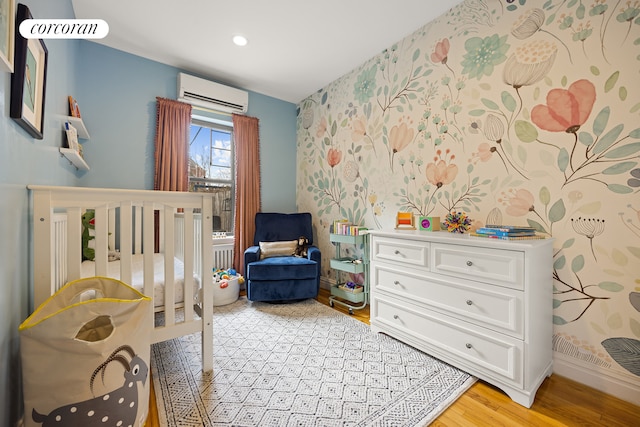 bedroom with a wall unit AC, light wood-type flooring, wallpapered walls, a nursery area, and baseboards
