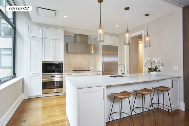 kitchen featuring wall chimney range hood, tasteful backsplash, a sink, a peninsula, and built in appliances