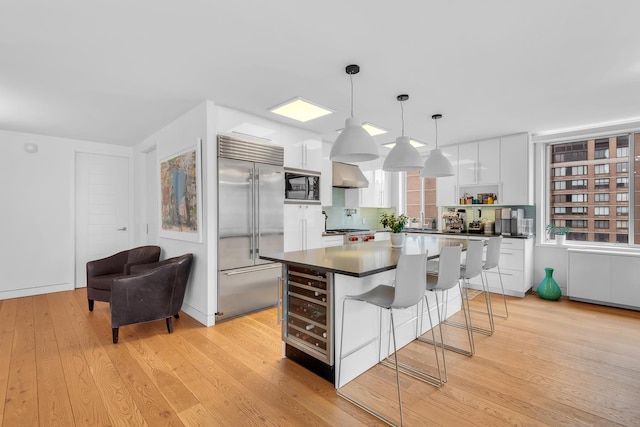 kitchen with built in appliances, dark countertops, a kitchen bar, light wood-type flooring, and wall chimney exhaust hood
